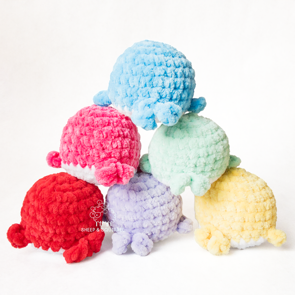 Six little amigurumi crochet stuffed animal whales in an assortment of colors stacked in a pyramid facing backward showing their tails