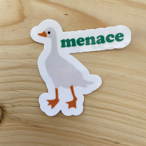 Die cut glossy vinyl sticker with a white goose that says "menace" on wood background