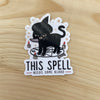 Die cut glossy vinyl sticker featuring a black cat with a witch hat, a knife in its mouth, on a seance circle that says "this spell needs some blood" on a wood background