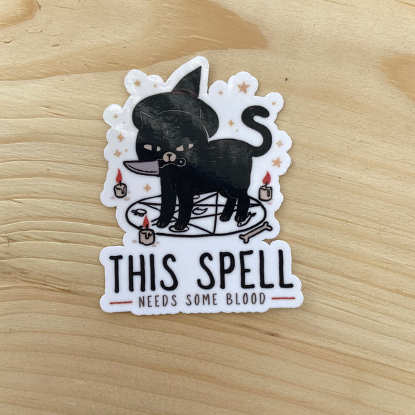 Die cut glossy vinyl sticker featuring a black cat with a witch hat, a knife in its mouth, on a seance circle that says "this spell needs some blood" on a wood background