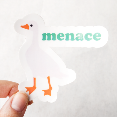 Die cut glossy vinyl sticker with a white goose that says "menace" held between tips of thumb and forefinger