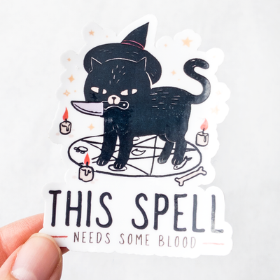 Die cut glossy vinyl sticker featuring a black cat with a witch hat, a knife in its mouth, on a seance circle that says "this spell needs some blood" being held between tips of thumb and forefinger