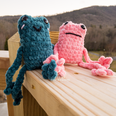 Two crochet amigurumi leggy frogs sitting on railing outside